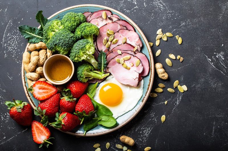 Teller mit einer bunten Auswahl an Speisen: Schinken mit Kürbiskernen, Brokkoli, ein Spiegelei mit flüssigem Eigelb, frische Erdbeeren, Erdnüsse in der Schale und eine kleine Schale mit Sauce oder Dressing. Der Teller steht auf dunklem Untergrund, umgeben von verstreuten Kürbiskernen und Erdnüssen.