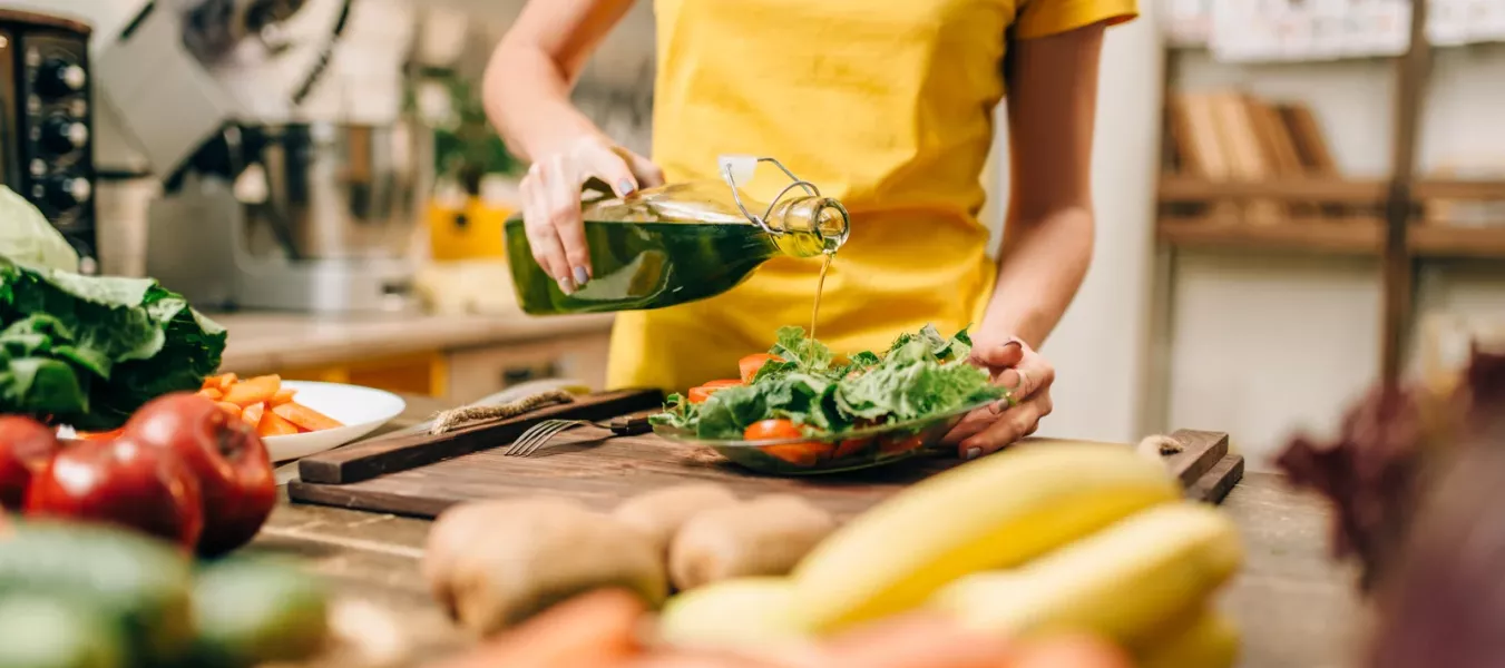 Person in gelbem Shirt gießt Olivenöl über einen frischen Salat in einer Küche. Auf der Arbeitsfläche liegen verschiedene Gemüse wie Karotten, Tomaten und Bananen. Im Hintergrund sind Küchenregale mit Gläsern und Behältern sowie Haushaltsgeräte zu sehen.