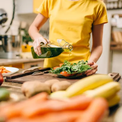Person in gelbem Shirt gießt Olivenöl über einen frischen Salat in einer Küche. Auf der Arbeitsfläche liegen verschiedene Gemüse wie Karotten, Tomaten und Bananen. Im Hintergrund sind Küchenregale mit Gläsern und Behältern sowie Haushaltsgeräte zu sehen.