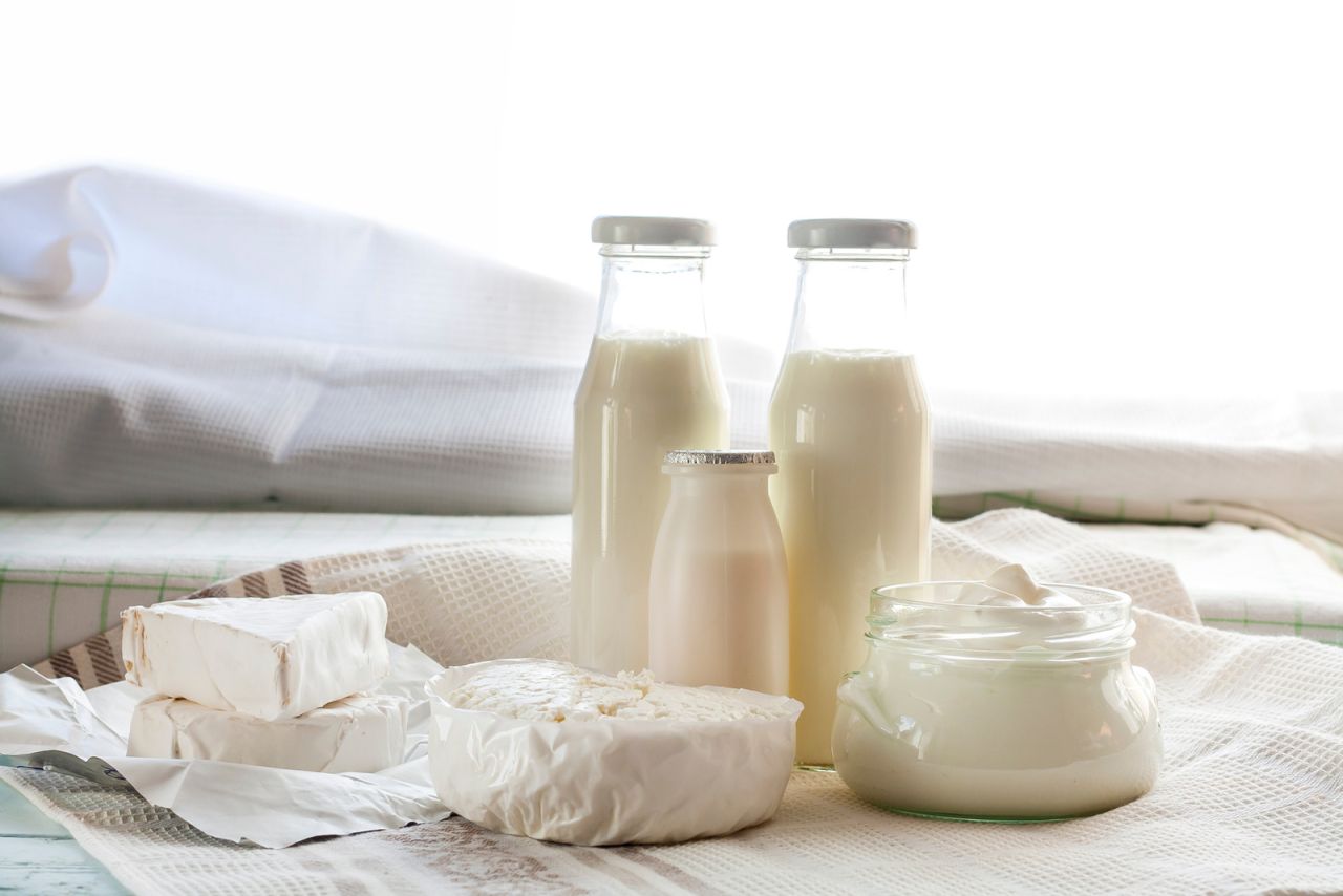 Verschiedene Milchprodukte auf einem mit Stoff bedeckten Untergrund: zwei große und eine kleine Glasflasche mit Milch, ein Glas mit Joghurt oder Sahne sowie drei Käsesorten – zwei rechteckige Stücke in weißem Papier und ein teilweise ausgepackter runder Käse.