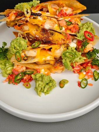 Ein Teller mit gestapelten Quesadillas, gefüllt mit Garnelen, geschmolzenem Käse und weiteren Zutaten. Die Quesadillas sind garniert mit Guacamole, gehackten Tomaten, grünen und roten Chilistreifen sowie frischer Petersilie.