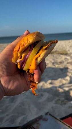 Eine Hand hält einen Burger mit geschmolzenem Käse, roten Zwiebelringen und einer gegrillten Ananasscheibe. Im Hintergrund sind ein Sandstrand und das Meer unter klarem, blauem Himmel zu sehen.