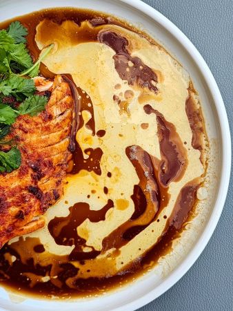 Nahaufnahme eines gebratenen Lachsfilets auf einem weißen Teller, garniert mit frischem Koriander. Rund um den Fisch sind kunstvoll helle und dunkle Soßen verteilt.