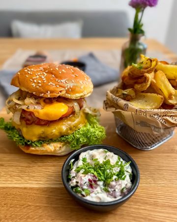 Nahaufnahme eines Cheeseburgers mit Sesambrötchen, Salat, Tomate, Käse und Zwiebeln, serviert mit Kartoffelwedges in einem kleinen Metallkorb mit Papierauslage und einem Schälchen Dip, garniert mit Kräutern und roten Zwiebeln.