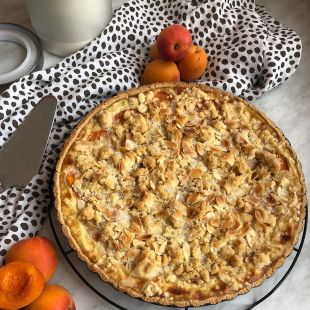 Frisch gebackene Aprikosentarte mit krümeligem Mandel-Zucker-Topping auf einem schwarzen Kuchengitter. Um die Tarte liegen ganze und halbierte Aprikosen. Eine weiße Stoffserviette mit schwarzen Punkten ist dekorativ drapiert, im Hintergrund steht ein Glas mit weißer Flüssigkeit.