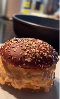 Nahaufnahme eines Burgers mit Sesambrötchen und geschmolzenem Käse, der an den Seiten herausläuft. Im Hintergrund sind eine schwarze Pfanne und unscharfe Küchenutensilien zu sehen.