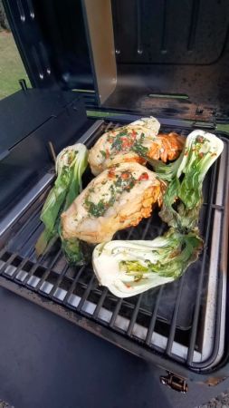 Geöffneter Grill mit drei gewürzten Hummerstücken und vier halbierten, ebenfalls gewürzten Pak-Choi-Stücken. Rauch steigt sichtbar vom Grill auf, während die Zutaten gegart werden.