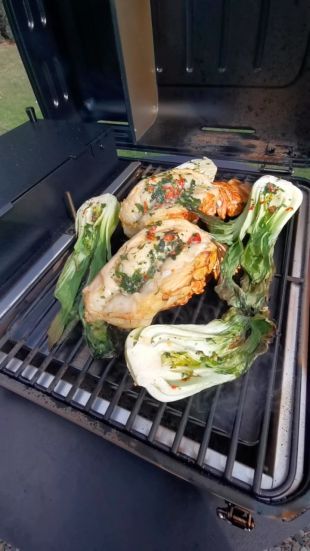 Geöffneter Grill mit drei gewürzten Hummerstücken und vier halbierten, ebenfalls gewürzten Pak-Choi-Stücken. Rauch steigt sichtbar vom Grill auf, während die Zutaten gegart werden.