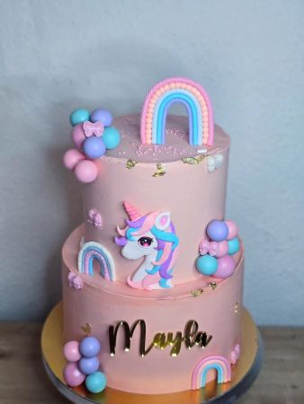 Zweistöckige rosa Torte mit pastellfarbenen Dekorationen. Die obere Etage zeigt ein Regenbogenmotiv, rosa Streusel und Kugeln in Pastellfarben. Die untere Etage ist mit einem weiteren Regenbogen, einem Einhornkopf mit pinkem Horn und bunter Mähne sowie dem Namen ‚Mayla‘ in goldenen Buchstaben verziert. Goldene Blättchen und weitere pastellfarbene Kugeln schmücken beide Etagen.
