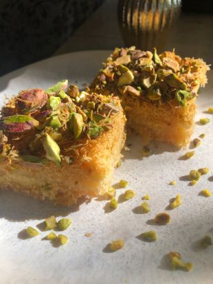 Zwei Stück Künefe, ein traditionelles Dessert aus dem Nahen Osten, aus feinen Teigfäden und süßem Sirup, serviert auf einem weißen Teller. Die Künefe ist mit gehackten Pistazien bestreut, weitere Pistazienstückchen liegen rund um das Gebäck.