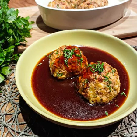 Käse-Laugenknödel in brauner Sauce, serviert in einer Schüssel und mit frischen Kräutern garniert; im Hintergrund weitere Knödel in einer zweiten Schale.