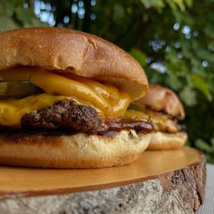 Zwei Cheeseburger mit Rindfleischpatty, geschmolzenem Cheddar, Gewürzgurken und Barbecuesauce auf einem Holzbrett, im Hintergrund grüne Pflanzen.