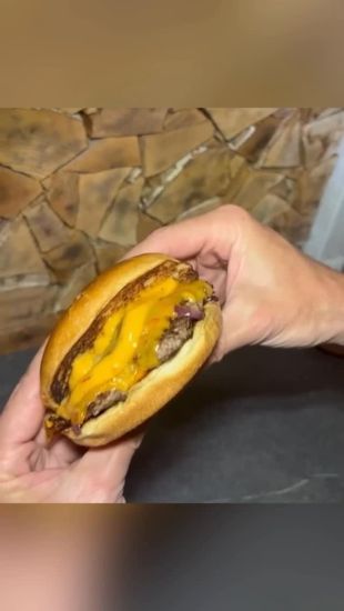 Cheeseburger mit Rindfleischpatty, geschmolzenem Cheddar und roten Zwiebeln, gehalten von zwei Händen vor einer Steinwand.