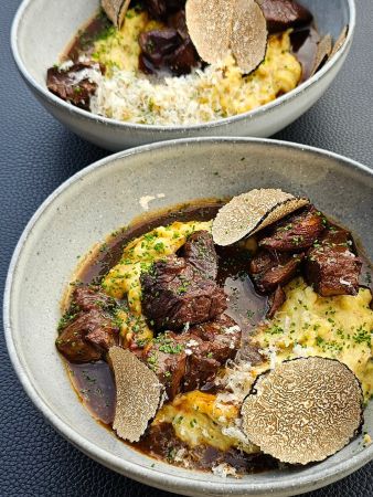 Zwei Teller mit geschmortem Rindfleisch auf cremigem Kartoffelpüree, garniert mit Trüffelscheiben und frischen Kräutern.