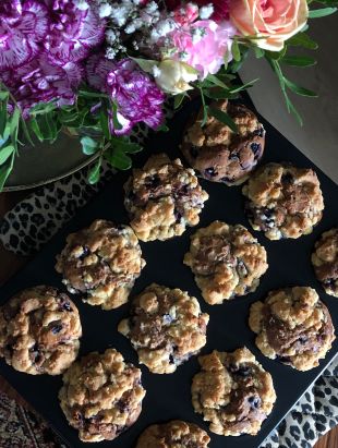 Wildheidelbeer-Muffins mit goldbrauner Kruste, sichtbaren Beeren und knusprigen Streuseln, dekorativ angerichtet unter einem bunten Blumenstrauß.