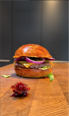 Nahaufnahme eines Gourmet-Burgers auf einer Holzoberfläche: mit glänzendem, getoastetem Brötchen, Rindfleischpatty, geschmolzenem Käse, roten Zwiebeln, Gewürzgurken und frischem Salat.