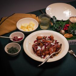 Festlich gedeckter Tisch mit einem Hauptgericht aus gerösteten Möhren in brauner Butter, garniert mit zerbröseltem Feta und Granatapfelkernen. Um das Gericht stehen kleine Schalen mit Butter, Kräutern und weiteren Zutaten.