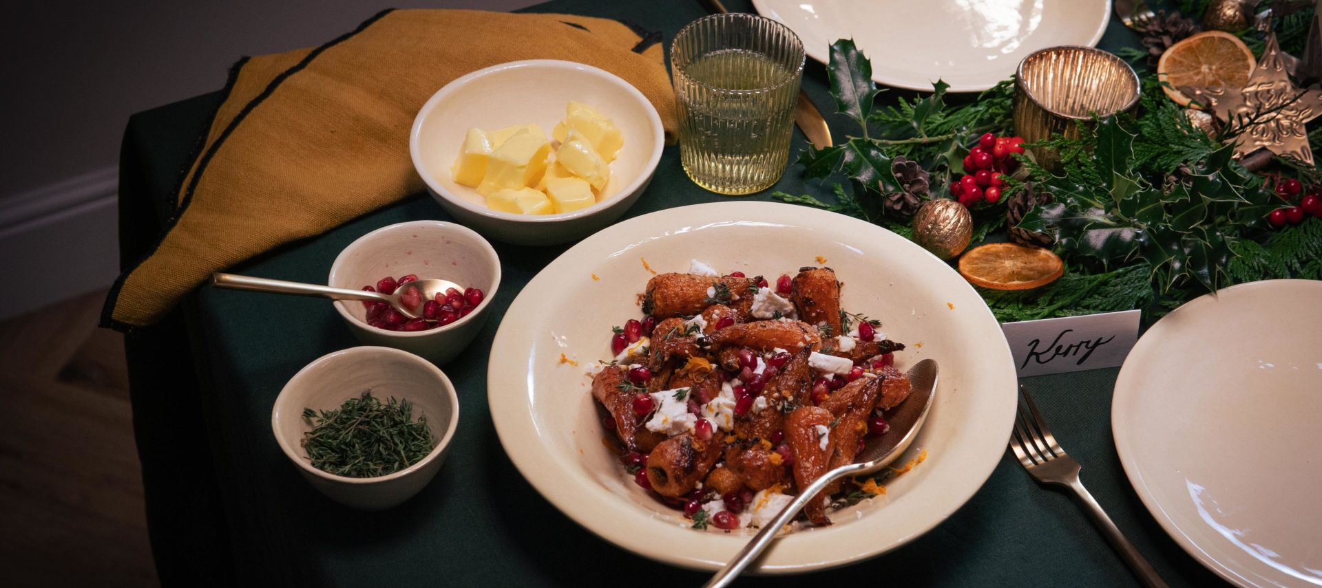 Festlich gedeckter Tisch mit einem Hauptgericht aus gerösteten Möhren in brauner Butter, garniert mit zerbröseltem Feta und Granatapfelkernen. Um das Gericht stehen kleine Schalen mit Butter, Kräutern und weiteren Zutaten.