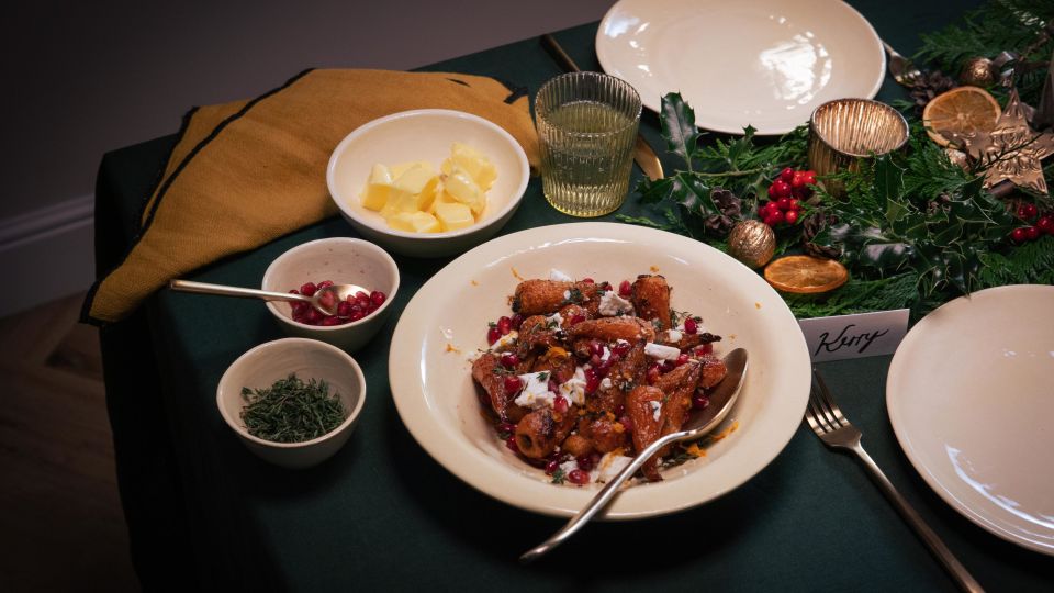 Festlich gedeckter Tisch mit einem Hauptgericht aus gerösteten Möhren in brauner Butter, garniert mit zerbröseltem Feta und Granatapfelkernen. Um das Gericht stehen kleine Schalen mit Butter, Kräutern und weiteren Zutaten.