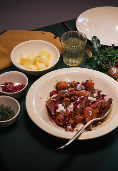Festlich gedeckter Tisch mit einem Hauptgericht aus gerösteten Möhren in brauner Butter, garniert mit zerbröseltem Feta und Granatapfelkernen. Um das Gericht stehen kleine Schalen mit Butter, Kräutern und weiteren Zutaten.