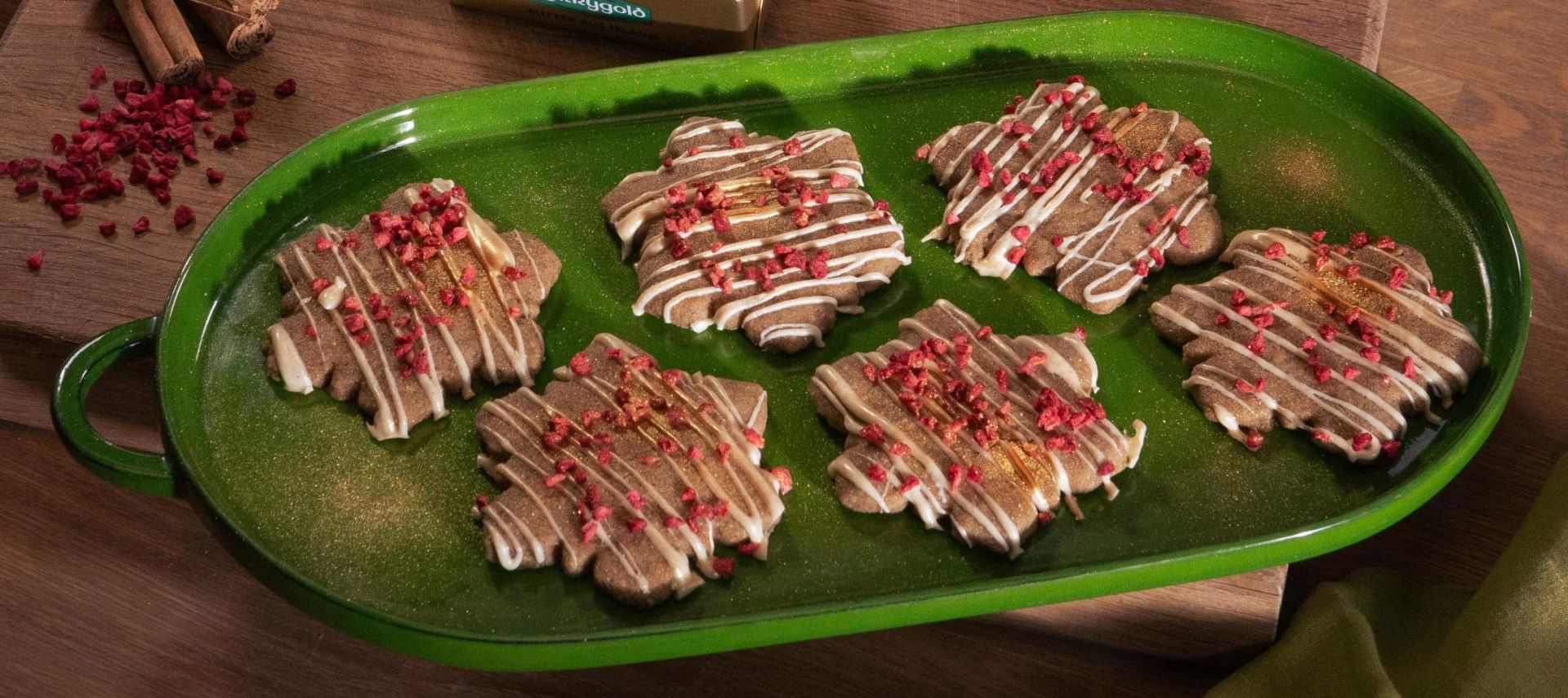 Festlich gedeckter Holztisch mit einem grünen Tablett voller Schneeflocken-förmigem Shortbread, verziert mit weißem Zuckerguss und gefriergetrockneten Himbeerstückchen. Daneben zwei Gläser Milch und ein Holzbrett mit einem Stück Kerrygold Butter. Im Hintergrund eine Schale mit Orangen, Tannenzapfen und grüner Dekoration.
