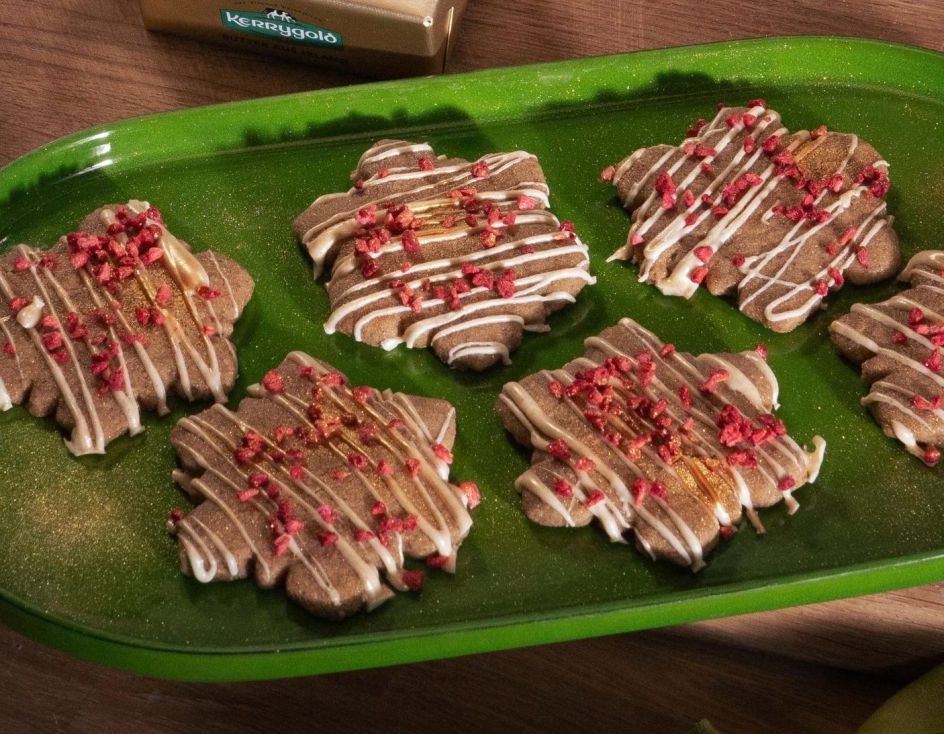 Festlich gedeckter Holztisch mit einem grünen Tablett voller Schneeflocken-förmigem Shortbread, verziert mit weißem Zuckerguss und gefriergetrockneten Himbeerstückchen. Daneben zwei Gläser Milch und ein Holzbrett mit einem Stück Kerrygold Butter. Im Hintergrund eine Schale mit Orangen, Tannenzapfen und grüner Dekoration.
