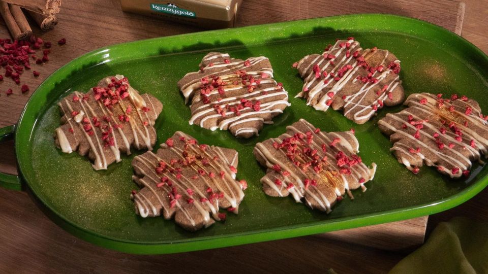 Festlich gedeckter Holztisch mit einem grünen Tablett voller Schneeflocken-förmigem Shortbread, verziert mit weißem Zuckerguss und gefriergetrockneten Himbeerstückchen. Daneben zwei Gläser Milch und ein Holzbrett mit einem Stück Kerrygold Butter. Im Hintergrund eine Schale mit Orangen, Tannenzapfen und grüner Dekoration.