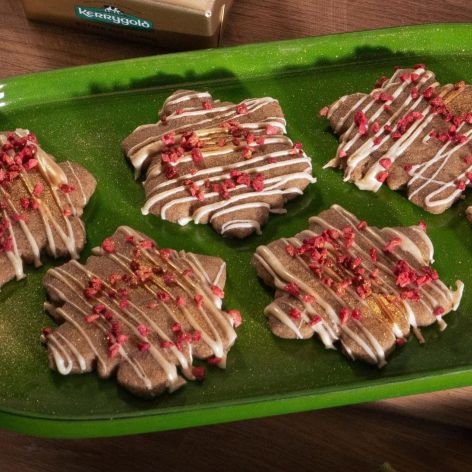Festlich gedeckter Holztisch mit einem grünen Tablett voller Schneeflocken-förmigem Shortbread, verziert mit weißem Zuckerguss und gefriergetrockneten Himbeerstückchen. Daneben zwei Gläser Milch und ein Holzbrett mit einem Stück Kerrygold Butter. Im Hintergrund eine Schale mit Orangen, Tannenzapfen und grüner Dekoration.