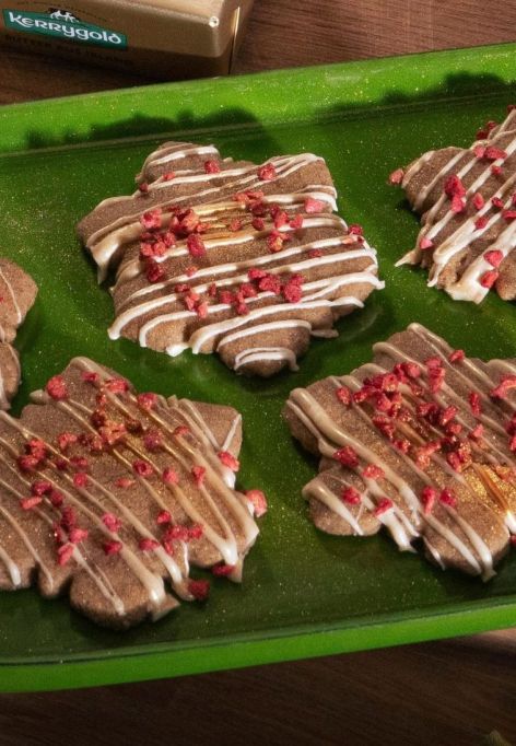 Festlich gedeckter Holztisch mit einem grünen Tablett voller Schneeflocken-förmigem Shortbread, verziert mit weißem Zuckerguss und gefriergetrockneten Himbeerstückchen. Daneben zwei Gläser Milch und ein Holzbrett mit einem Stück Kerrygold Butter. Im Hintergrund eine Schale mit Orangen, Tannenzapfen und grüner Dekoration.