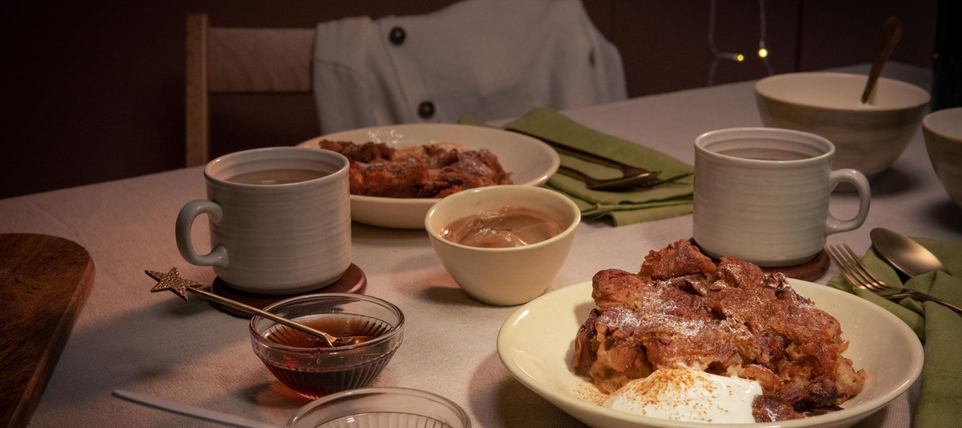 Gemütlich gedeckter Tisch mit zwei Tellern Croissant-Auflauf, jeweils serviert mit einem Klecks Crème fraîche. Dazu zwei Tassen Heißgetränk, kleine Schälchen mit Sauce und grüne Stoffservietten.