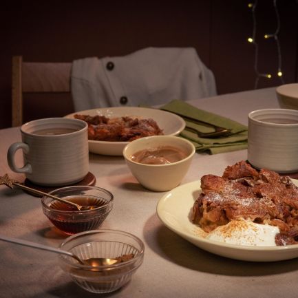 Gemütlich gedeckter Tisch mit zwei Tellern Croissant-Auflauf, jeweils serviert mit einem Klecks Crème fraîche. Dazu zwei Tassen Heißgetränk, kleine Schälchen mit Sauce und grüne Stoffservietten.