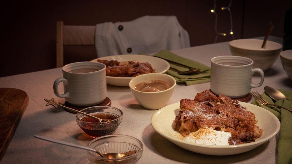 Gemütlich gedeckter Tisch mit zwei Tellern Croissant-Auflauf, jeweils serviert mit einem Klecks Crème fraîche. Dazu zwei Tassen Heißgetränk, kleine Schälchen mit Sauce und grüne Stoffservietten.