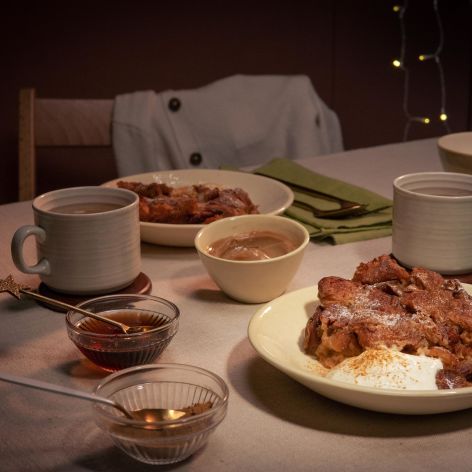 Gemütlich gedeckter Tisch mit zwei Tellern Croissant-Auflauf, jeweils serviert mit einem Klecks Crème fraîche. Dazu zwei Tassen Heißgetränk, kleine Schälchen mit Sauce und grüne Stoffservietten.