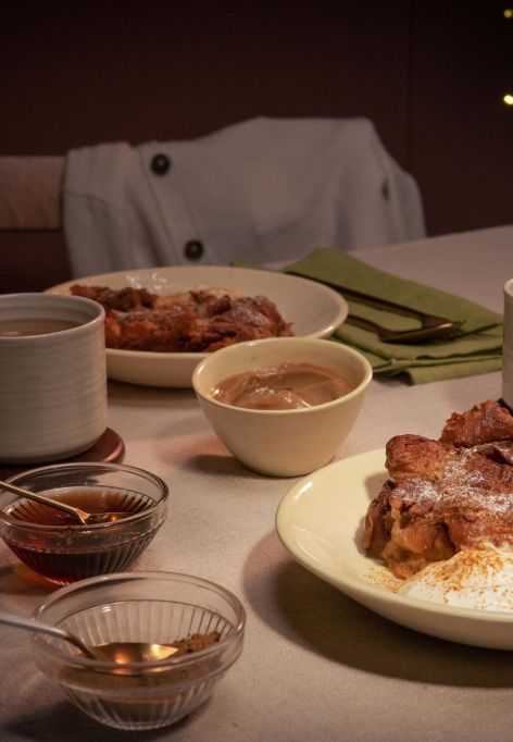 Gemütlich gedeckter Tisch mit zwei Tellern Croissant-Auflauf, jeweils serviert mit einem Klecks Crème fraîche. Dazu zwei Tassen Heißgetränk, kleine Schälchen mit Sauce und grüne Stoffservietten.