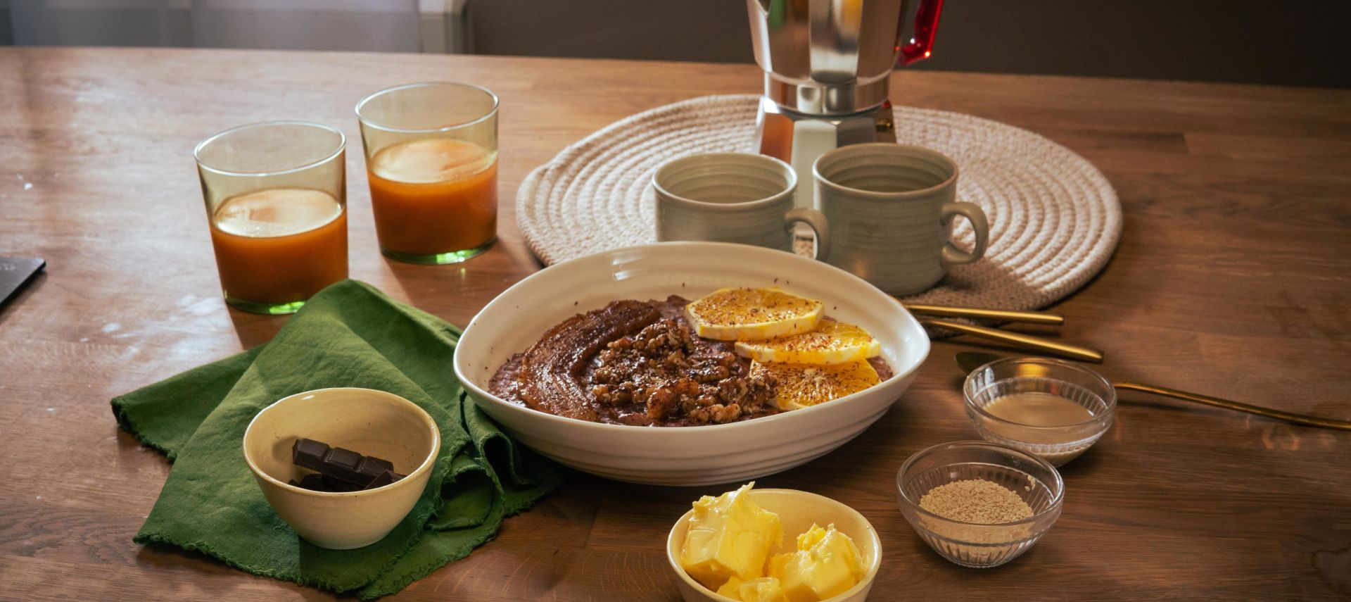 Frühstückstisch mit einer Schüssel Schoko-Tahini-Porridge, garniert mit Orangenscheiben. Drumherum kleine Schalen mit Zutaten wie Butter, braunem Zucker und Schokolade. Zwei Gläser mit Orangensaft, zwei Keramikbecher und eine Espressokanne stehen im Hintergrund.