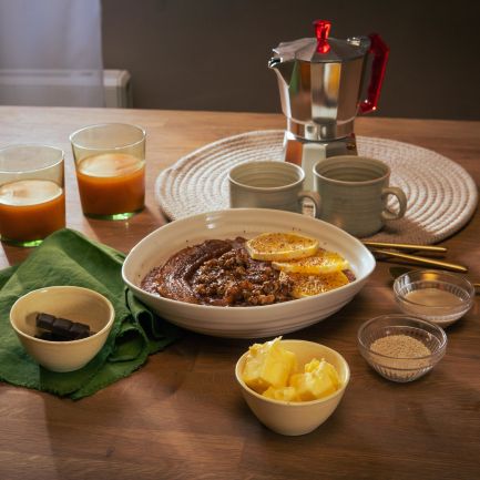 Frühstückstisch mit einer Schüssel Schoko-Tahini-Porridge, garniert mit Orangenscheiben. Drumherum kleine Schalen mit Zutaten wie Butter, braunem Zucker und Schokolade. Zwei Gläser mit Orangensaft, zwei Keramikbecher und eine Espressokanne stehen im Hintergrund.