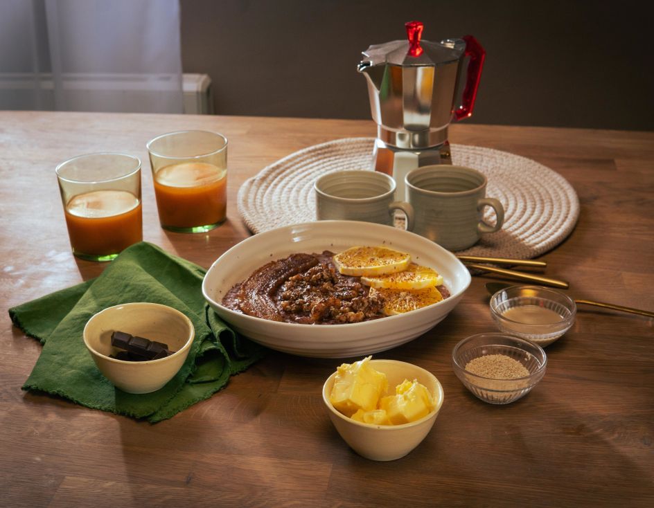 Frühstückstisch mit einer Schüssel Schoko-Tahini-Porridge, garniert mit Orangenscheiben. Drumherum kleine Schalen mit Zutaten wie Butter, braunem Zucker und Schokolade. Zwei Gläser mit Orangensaft, zwei Keramikbecher und eine Espressokanne stehen im Hintergrund.