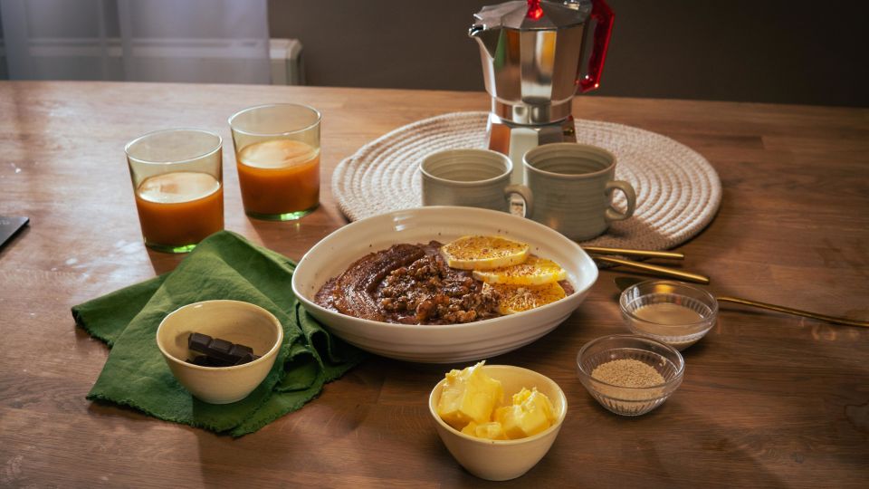Frühstückstisch mit einer Schüssel Schoko-Tahini-Porridge, garniert mit Orangenscheiben. Drumherum kleine Schalen mit Zutaten wie Butter, braunem Zucker und Schokolade. Zwei Gläser mit Orangensaft, zwei Keramikbecher und eine Espressokanne stehen im Hintergrund.