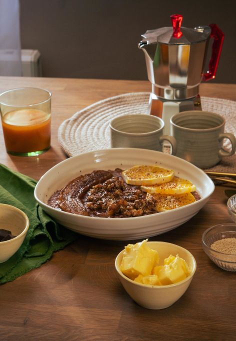 Frühstückstisch mit einer Schüssel Schoko-Tahini-Porridge, garniert mit Orangenscheiben. Drumherum kleine Schalen mit Zutaten wie Butter, braunem Zucker und Schokolade. Zwei Gläser mit Orangensaft, zwei Keramikbecher und eine Espressokanne stehen im Hintergrund.