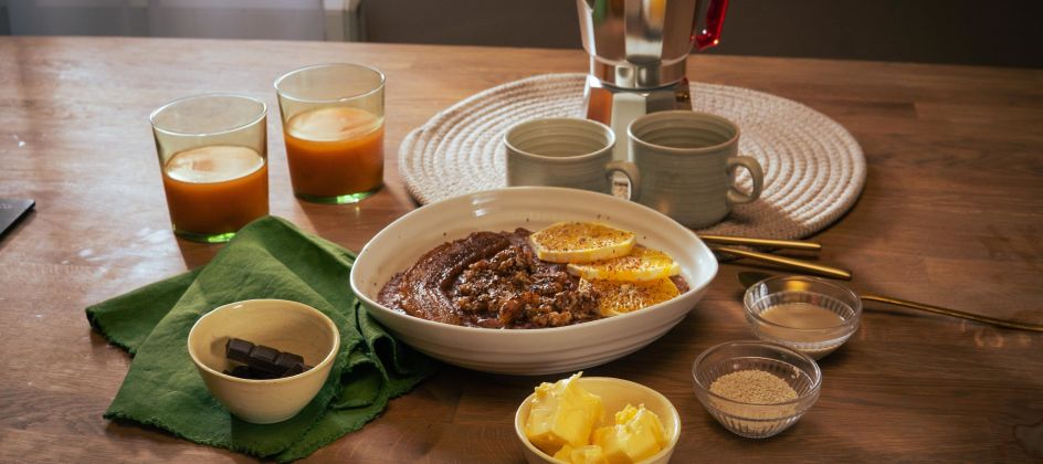 Frühstückstisch mit einer Schüssel Schoko-Tahini-Porridge, garniert mit Orangenscheiben. Drumherum kleine Schalen mit Zutaten wie Butter, braunem Zucker und Schokolade. Zwei Gläser mit Orangensaft, zwei Keramikbecher und eine Espressokanne stehen im Hintergrund.