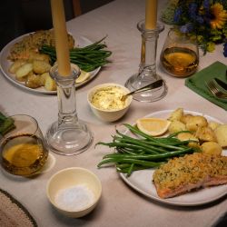 Festlich gedeckter Tisch mit zwei Tellern Kräuterbutter-Lachs, serviert mit grünen Bohnen und gerösteten Drillingkartoffeln. In der Mitte des Tisches stehen zwei brennende Stabkerzen in Glasständern.