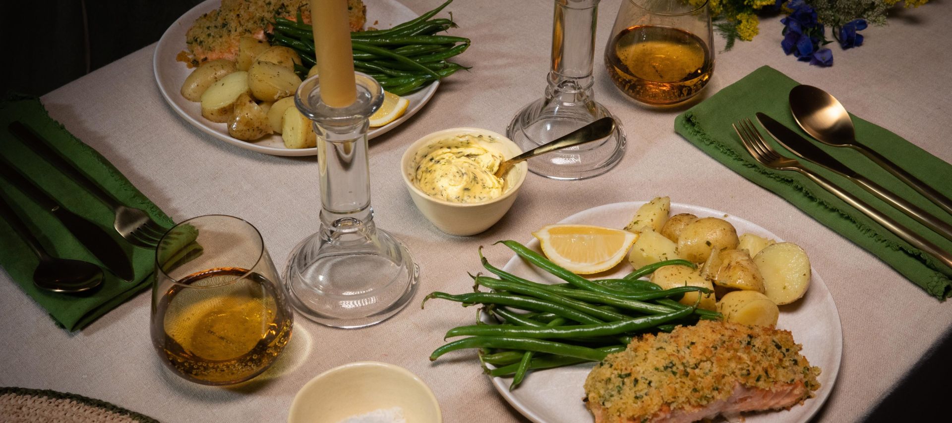 Festlich gedeckter Tisch mit zwei Tellern Kräuterbutter-Lachs, serviert mit grünen Bohnen und gerösteten Drillingkartoffeln. In der Mitte des Tisches stehen zwei brennende Stabkerzen in Glasständern.