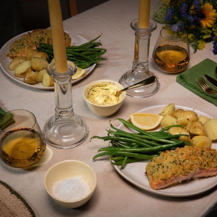 Festlich gedeckter Tisch mit zwei Tellern Kräuterbutter-Lachs, serviert mit grünen Bohnen und gerösteten Drillingkartoffeln. In der Mitte des Tisches stehen zwei brennende Stabkerzen in Glasständern.