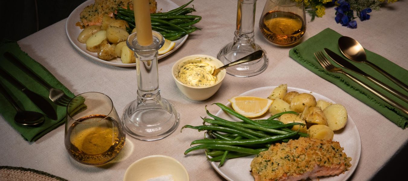 Festlich gedeckter Tisch mit zwei Tellern Kräuterbutter-Lachs, serviert mit grünen Bohnen und gerösteten Drillingkartoffeln. In der Mitte des Tisches stehen zwei brennende Stabkerzen in Glasständern.