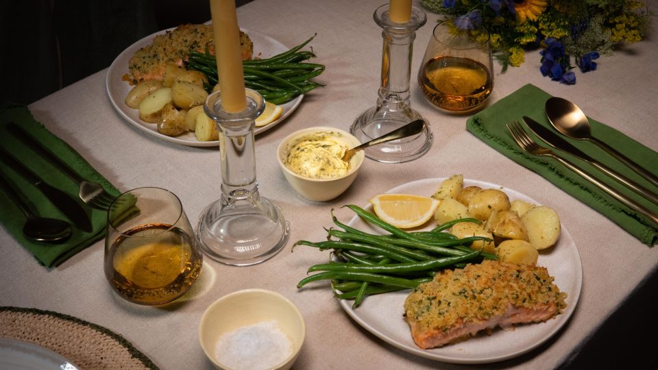 Festlich gedeckter Tisch mit zwei Tellern Kräuterbutter-Lachs, serviert mit grünen Bohnen und gerösteten Drillingkartoffeln. In der Mitte des Tisches stehen zwei brennende Stabkerzen in Glasständern.