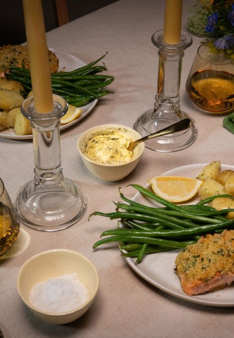 Festlich gedeckter Tisch mit zwei Tellern Kräuterbutter-Lachs, serviert mit grünen Bohnen und gerösteten Drillingkartoffeln. In der Mitte des Tisches stehen zwei brennende Stabkerzen in Glasständern.