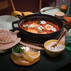 Tisch mit einer Pfanne Shakshuka – pochierte Eier in würziger Tomatensauce, garniert mit Kräutern und Klecksen von Frischkäse. Dazu Fladenbrot, Zitronenspalten und Kräuter.