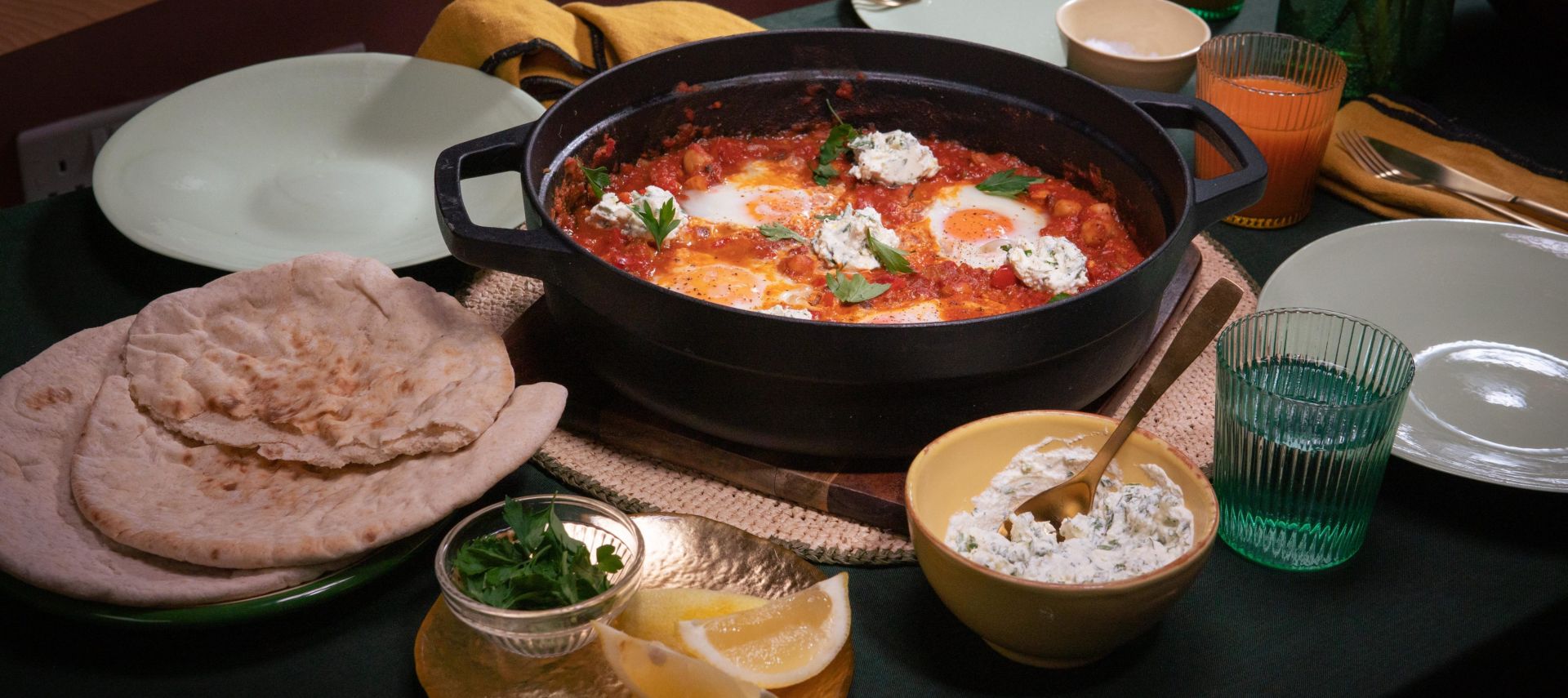 Tisch mit einer Pfanne Shakshuka – pochierte Eier in würziger Tomatensauce, garniert mit Kräutern und Klecksen von Frischkäse. Dazu Fladenbrot, Zitronenspalten und Kräuter.
