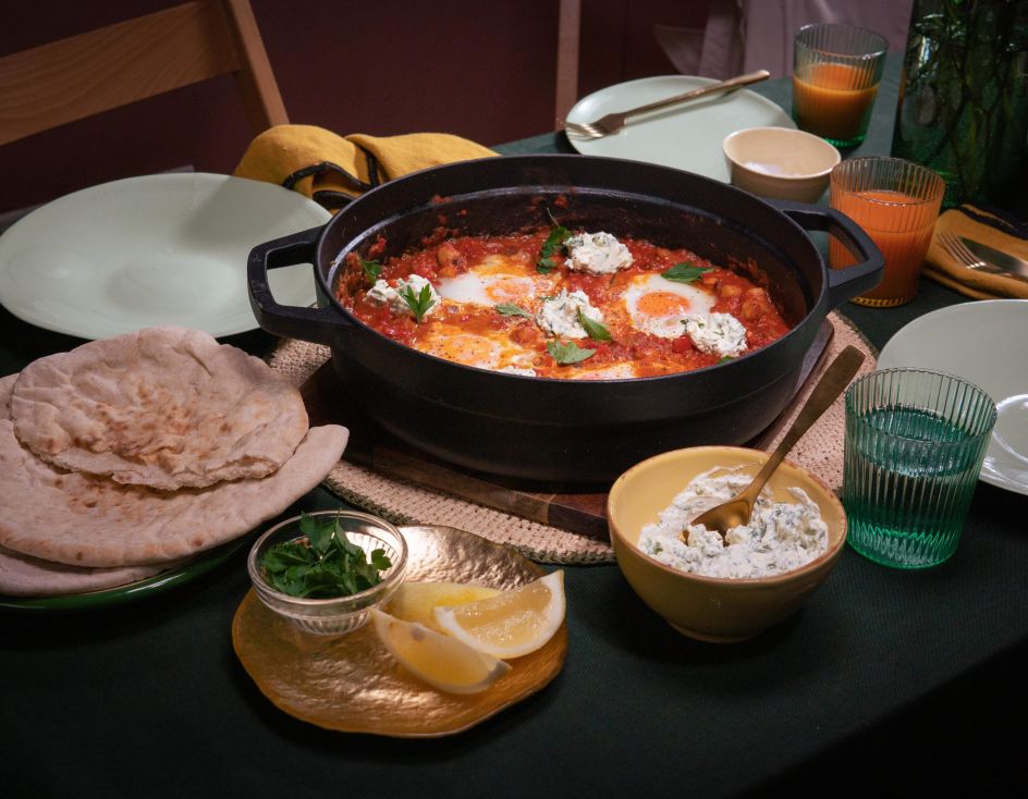 Tisch mit einer Pfanne Shakshuka – pochierte Eier in würziger Tomatensauce, garniert mit Kräutern und Klecksen von Frischkäse. Dazu Fladenbrot, Zitronenspalten und Kräuter.