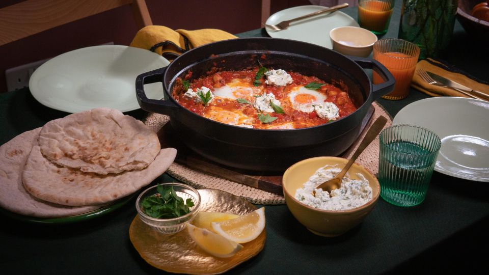 Tisch mit einer Pfanne Shakshuka – pochierte Eier in würziger Tomatensauce, garniert mit Kräutern und Klecksen von Frischkäse. Dazu Fladenbrot, Zitronenspalten und Kräuter.